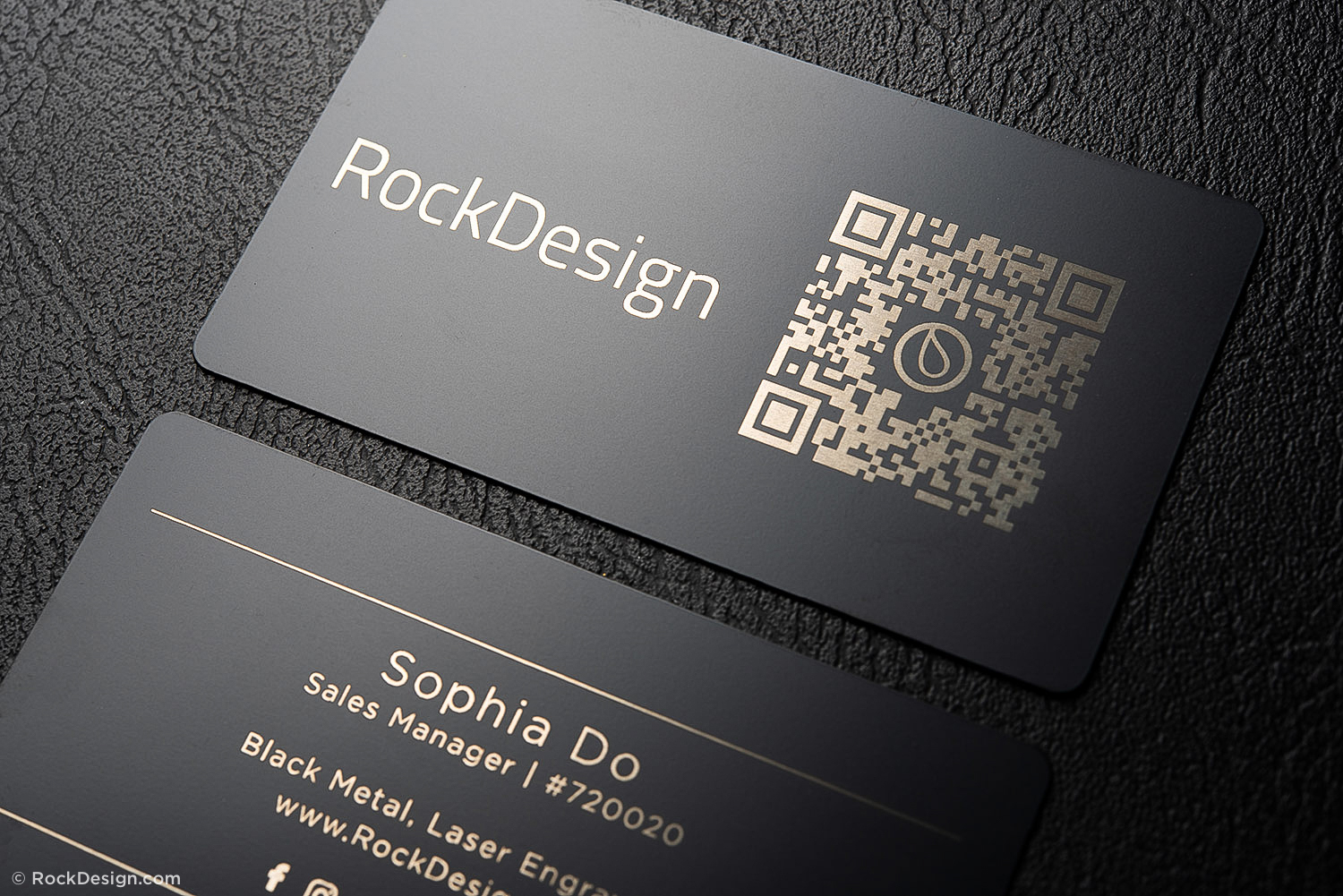 Premium black metal business cards from RockDesign featuring gold laser engraving, with company branding, QR code, and contact details displayed on a textured black surface.